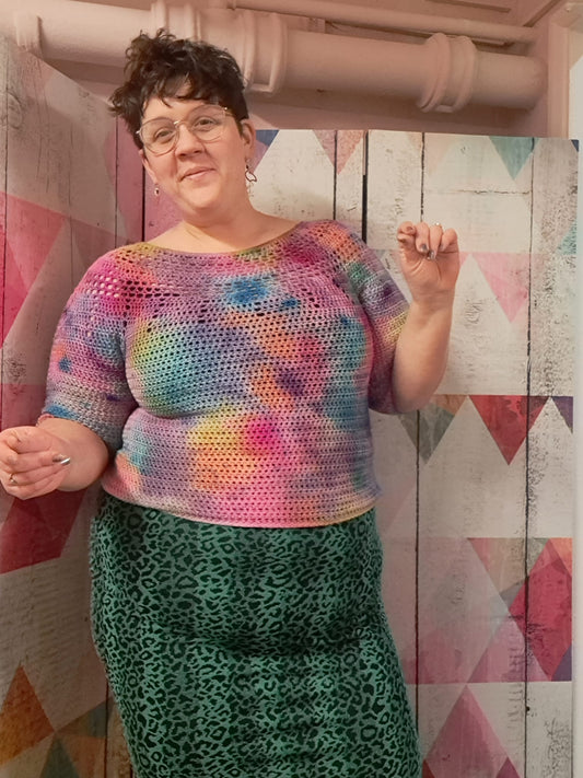 Eleanor, a fat, white woman smiles at the camera and does a sort of jig. She's wearing a tight, green, leopard-ish print skirt and a tie dyed effect, crochet jumper.