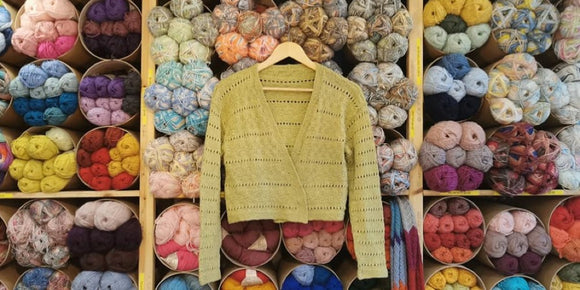 A hand knitted cardigan hangs infront of full shelves of yarn. They’re circular tubes set out kind of like honeycombs. The cardigan is green, has long sleeves and a simple texture and lace pattern. 