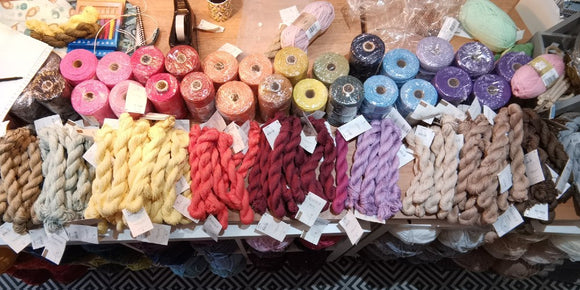 A shot taken from directly above our main table. On the table are piles of linen skeins and behind them big spools of macrame cotton in rough rainbow order. Behind that is just general shop detritus like coffee, pens, cellotape and recycled plastic bags. 
