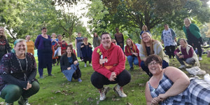 Eleanor and customers of Knit Nottingham take a socially distanced, timer selfie in the Aboretum. There are lots of people, mainly women, some crouching, some standing, some sitting all facing the camera. There’s lots of green trees and grass.