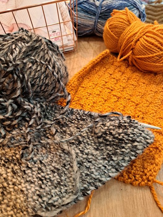 Two knitted swatches laid on a wooden table. The one at the front is greyish/blackish marled and the one underneath is a pumpkin orange in a basketweave variation. 
