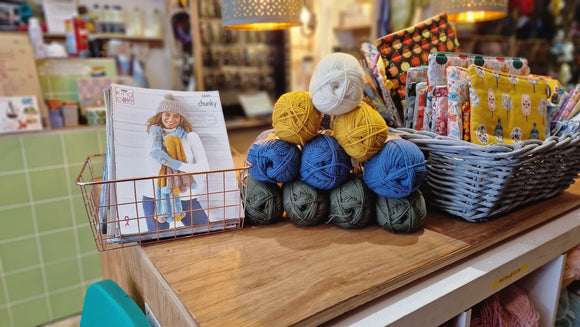 A pyramid of Wildwood Yarn on a wooden table with a knitting pattern featuring a scarf and a hat on the left. There's a basket of project bags on the right too. 