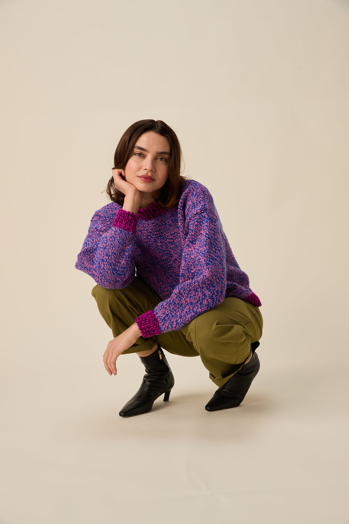 A model crouches in a studio setting wearing a purple and pink textured knit sweater, olive green pants, and black ankle boots against a solid beige background.