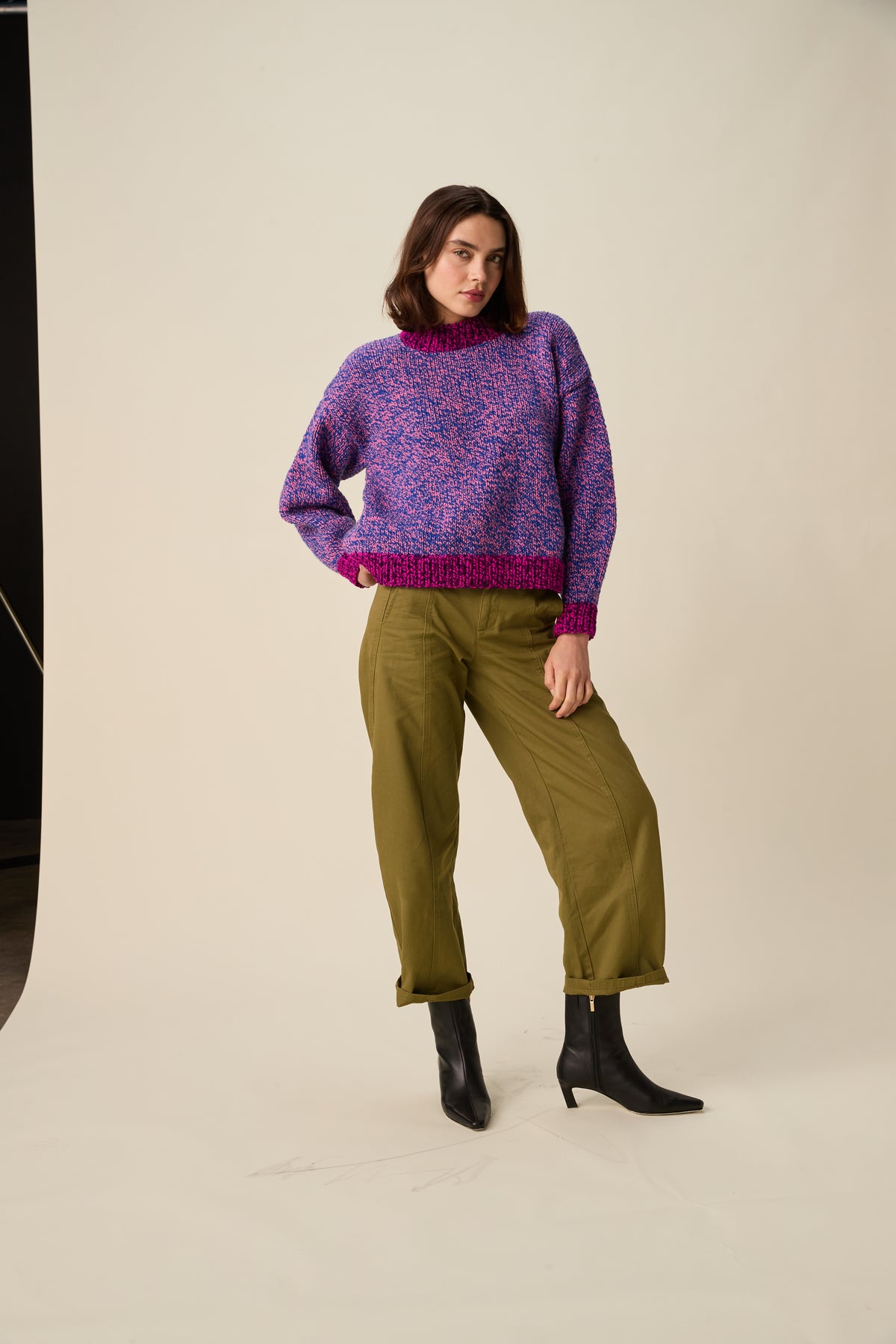 A woman modeling a purple and blue marled knit sweater with a magenta hem, paired with olive green trousers and black ankle boots, against a warm off-white studio background.