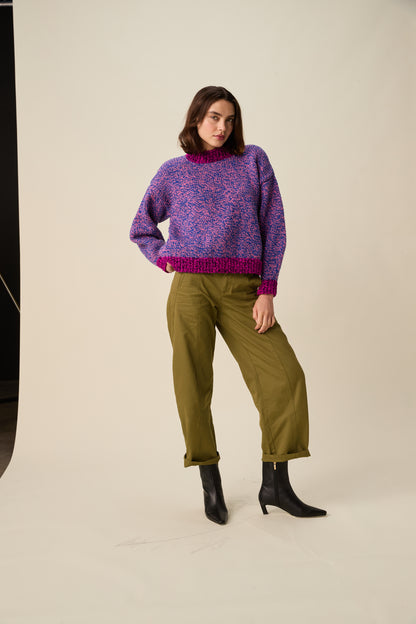 A woman modeling a purple and blue marled knit sweater with a magenta hem, paired with olive green trousers and black ankle boots, against a warm off-white studio background.