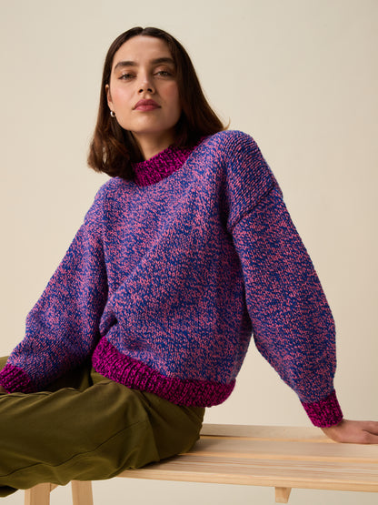 A woman wearing a chunky knit sweater with a purple and pink heathered pattern and magenta ribbed trim, seated on a light wood bench against a plain beige background.