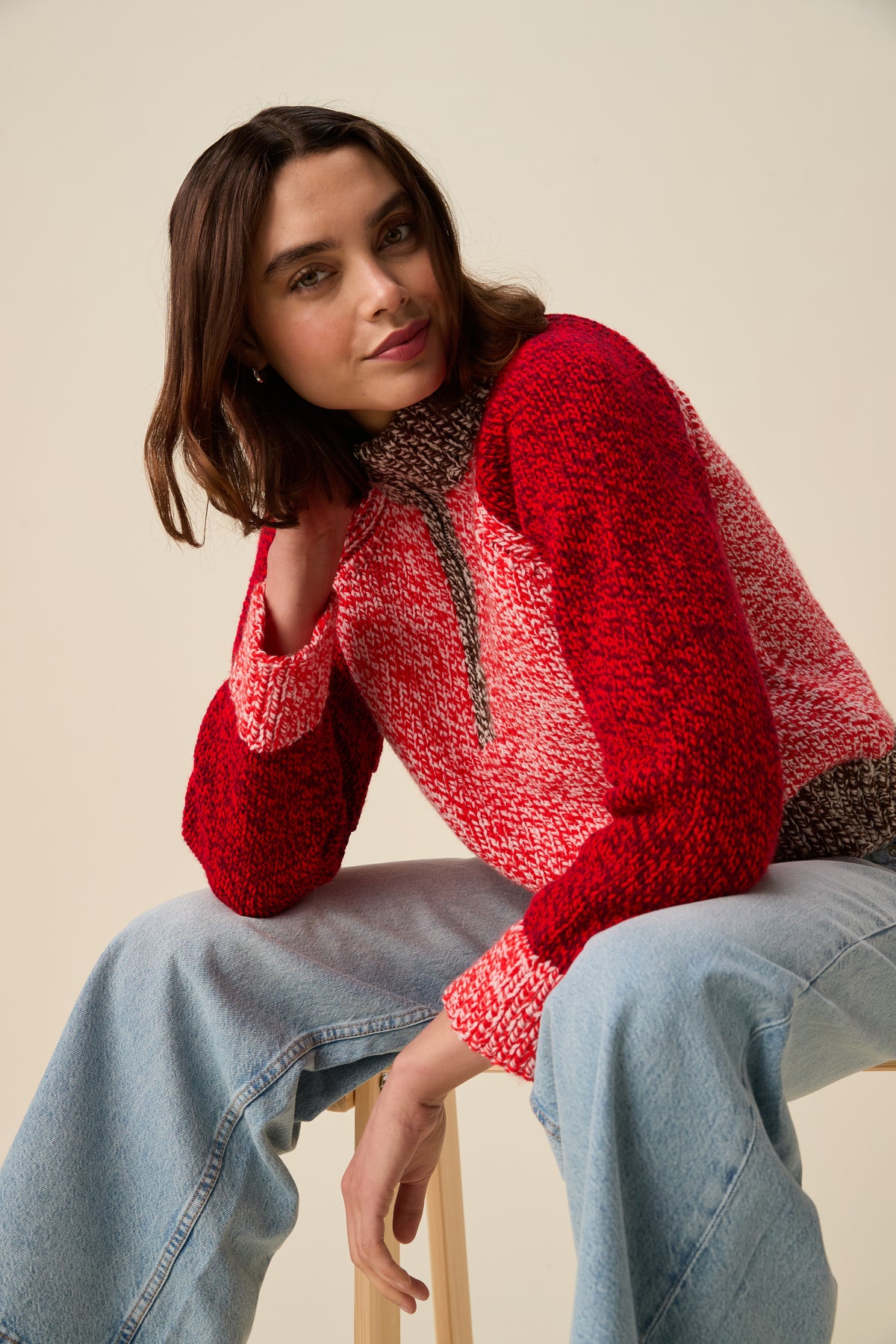 A woman wearing a red and white marled knit pullover sweater with brown textured trim, paired with light blue jeans, sitting on a wooden stool against a warm beige background.