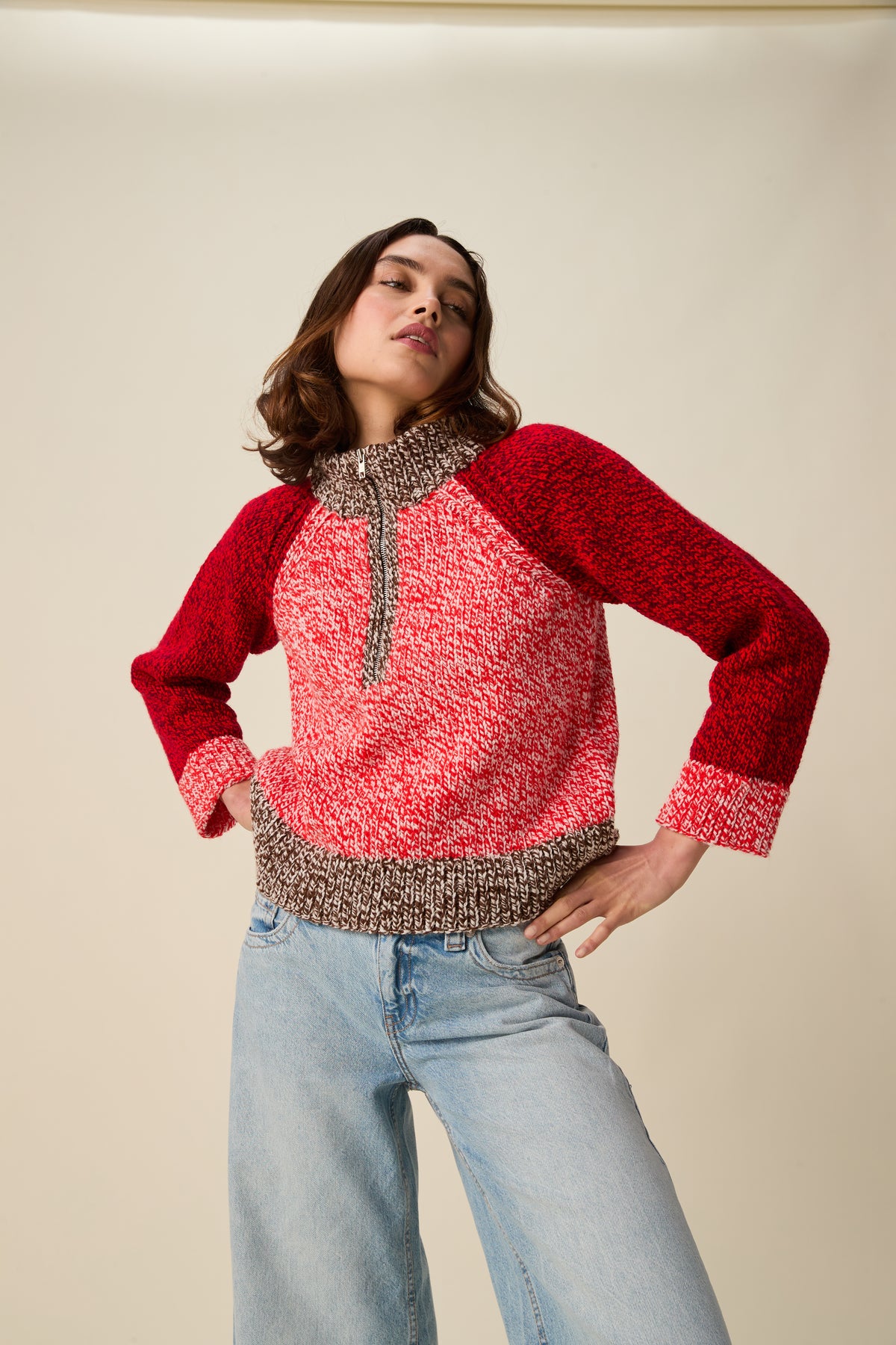 A woman modeling a marled knit half-zip sweater with red raglan sleeves and a pinkish-red body, paired with light-wash jeans against a plain beige studio background.