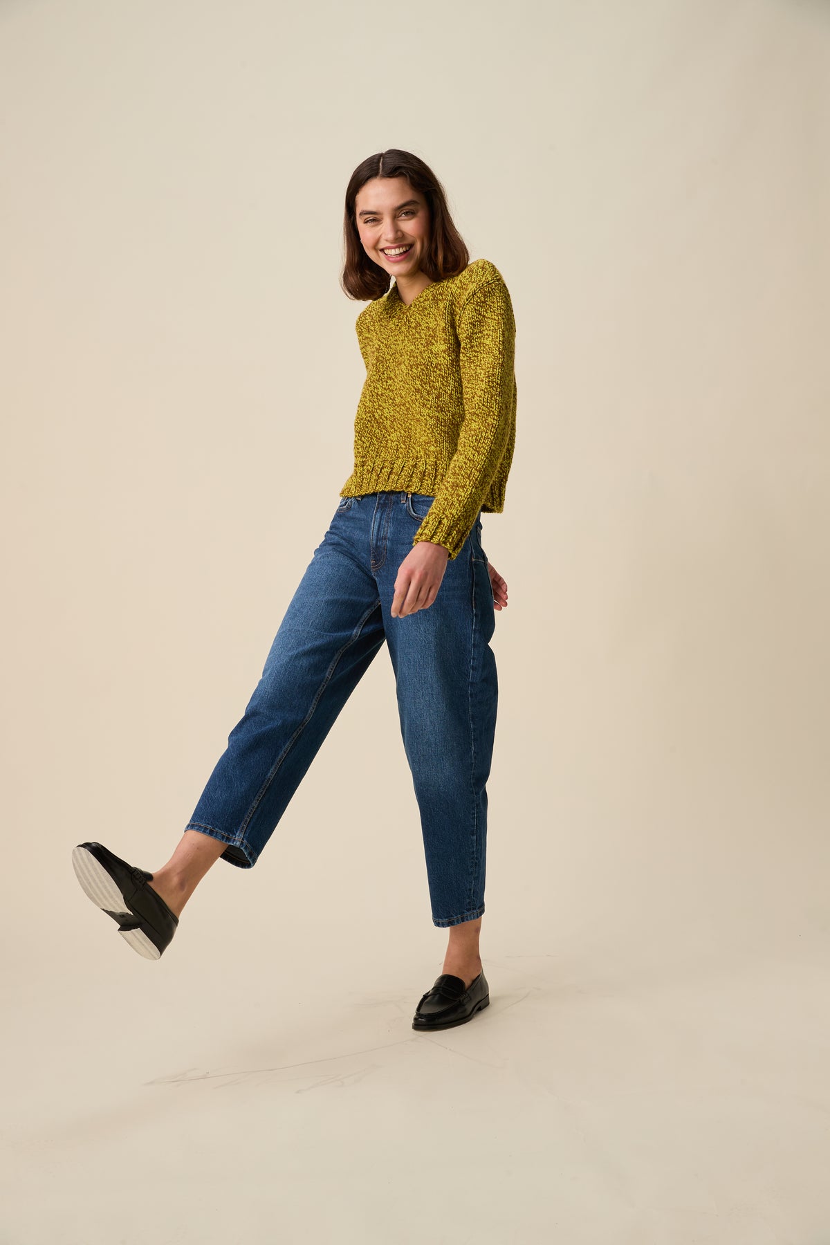 A woman modeling a textured green and brown knit sweater and blue cropped jeans with black loafers, standing against a solid beige studio background.