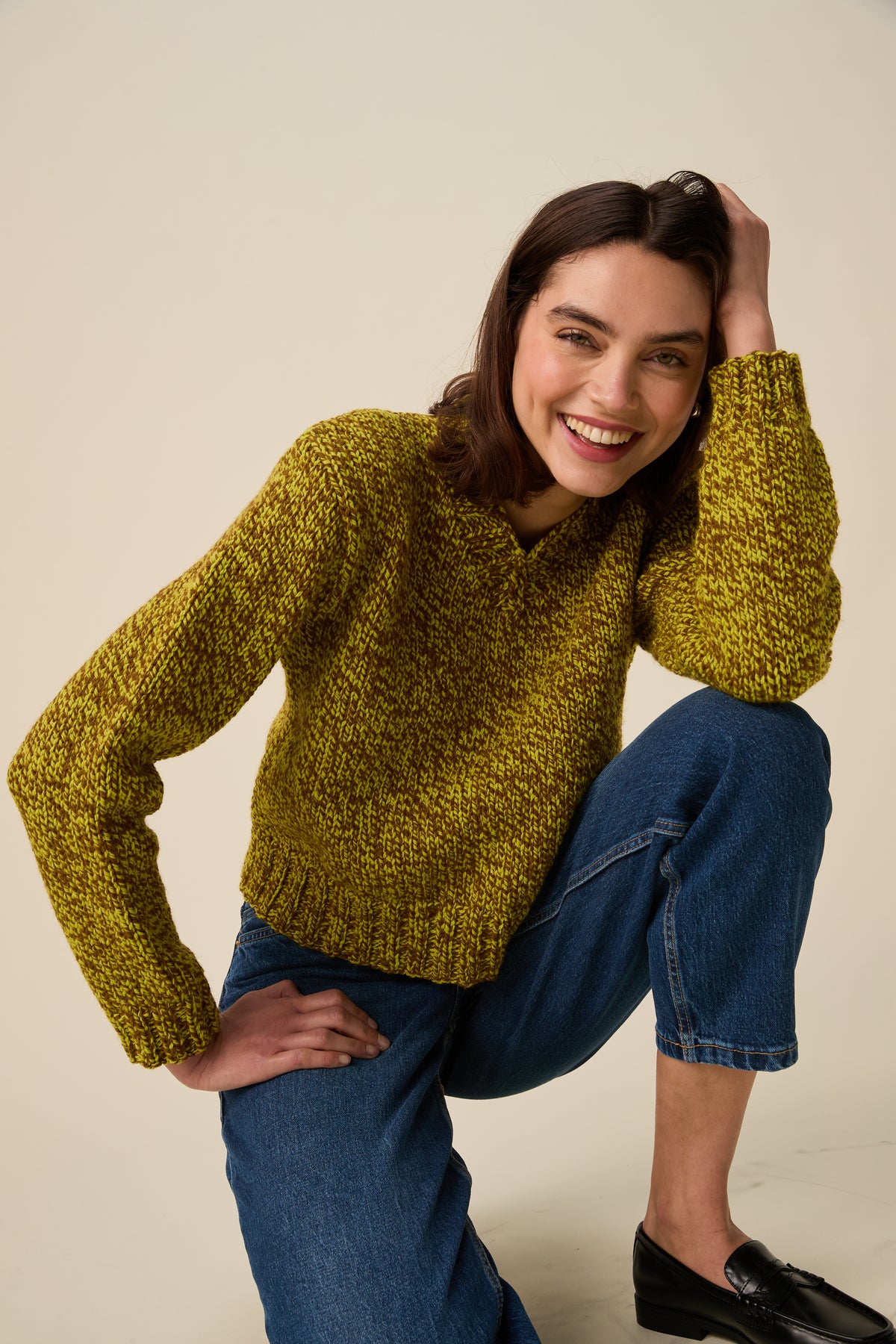 A woman wearing a chunky knit green and brown sweater and blue jeans, posing in a studio setting against a light beige background.