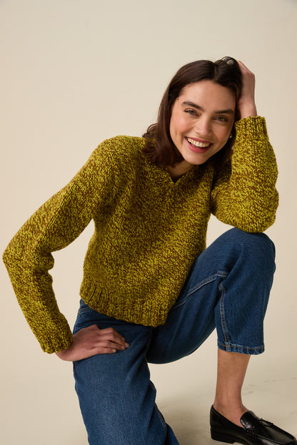 A woman wearing a chunky knit green and brown sweater and blue jeans, posing in a studio setting against a light beige background.